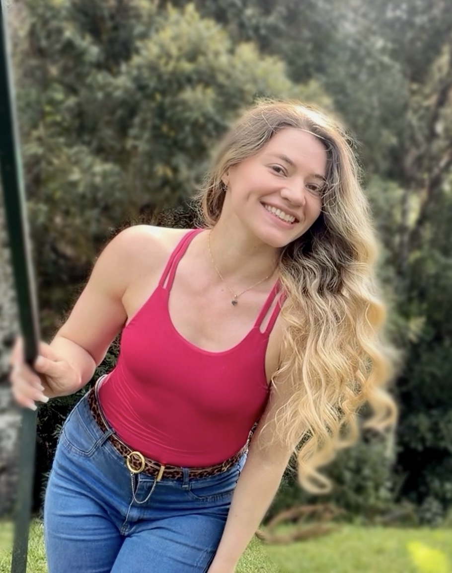 Emellyn Cracco sorrindo ao ar livre, em um parque, com regata rosa e calça jeans